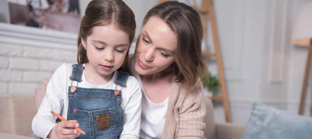 a moth kneels besides young daughter who is drawing with crayons in family home living room; Broward County Guardianship Lawyer concept image